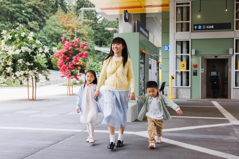 A mom and her two kids hold hands and walk near the entrance to South Bellevue Station on a sunny day