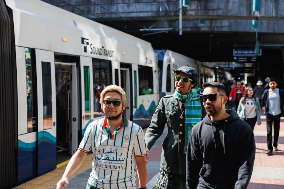 Three people in soccer jerseys walk along the sunny platform at International District Chinatown Station.