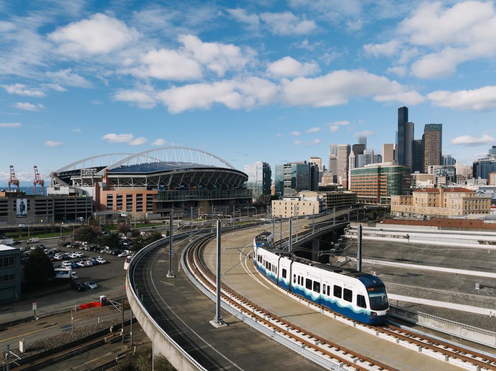 A two-car 2 Line train on the tracks with the stadium and downtown Seattle skyline in the background