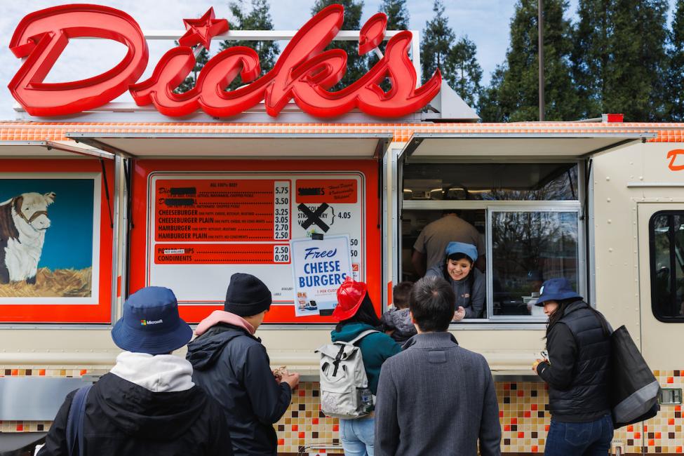 People line up at a food truck with a large red sign that reads "Dick's" on top of the truck