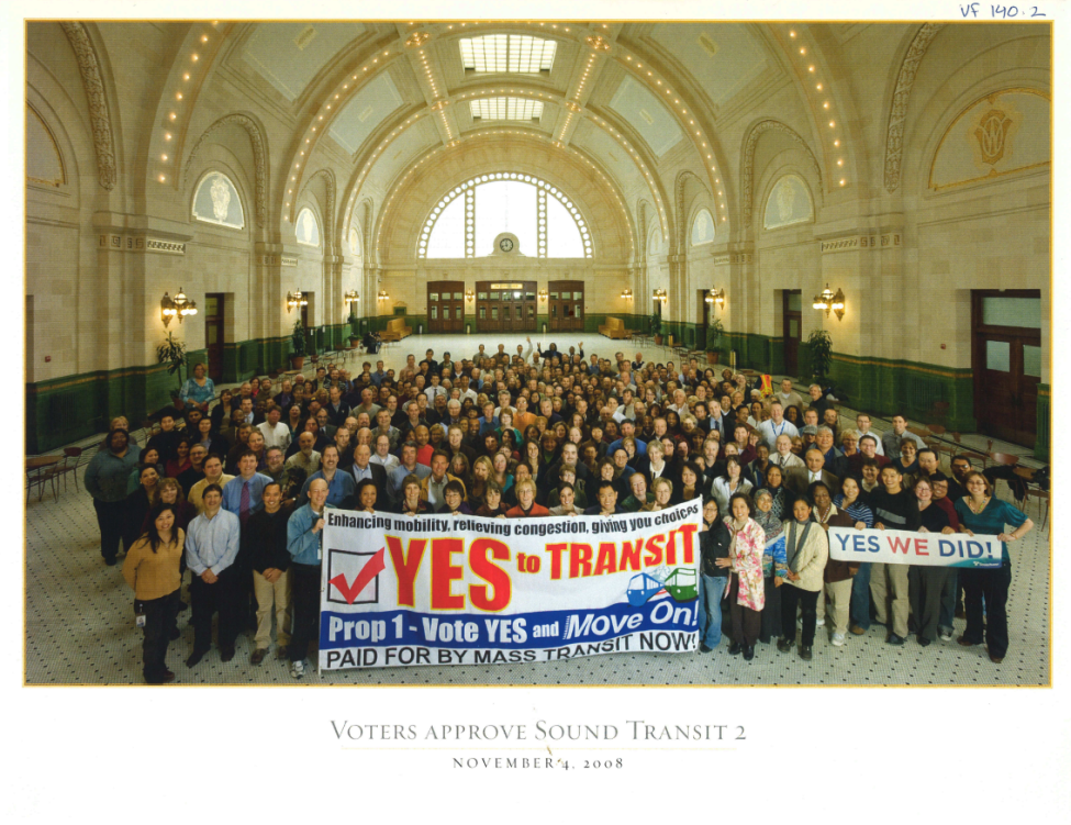 A large group of people gather in Union Station. Those in front hold a banner reading "Yes to transit"