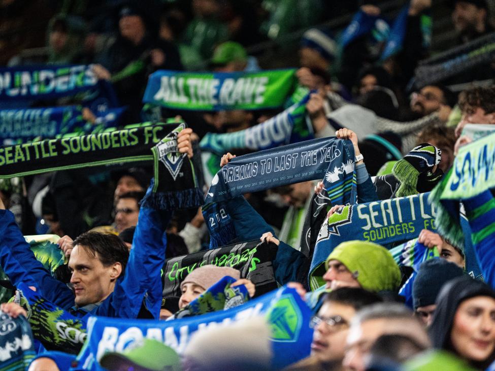 Fans dressed in green and blue hold their Seattle Sounders scarves up in the air at a game.