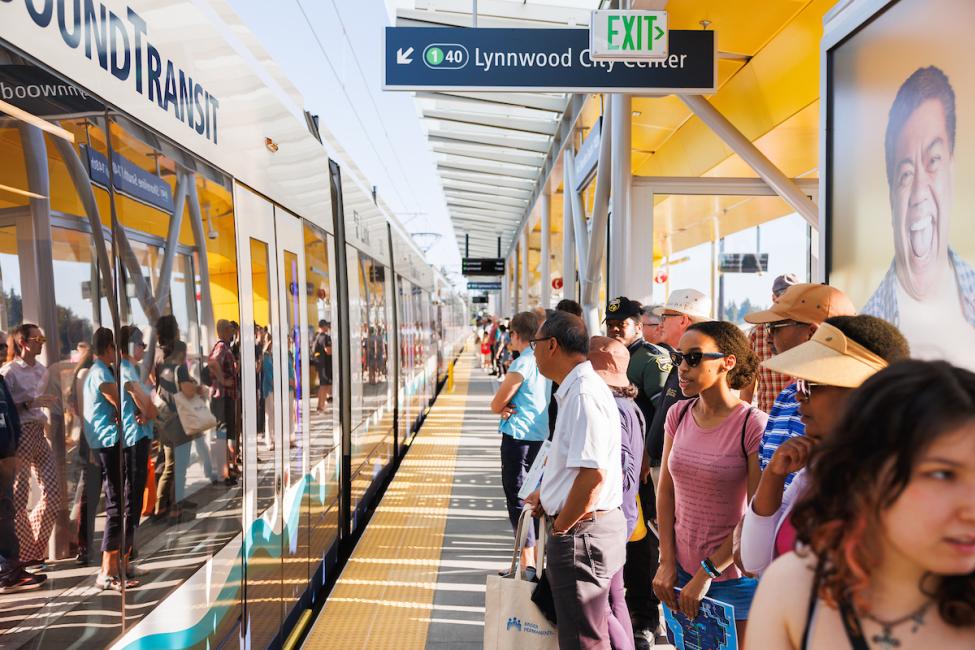 A large group of people wait behind the yellow line on the platform at Lynnwood City Center Station as a Link train pulls up