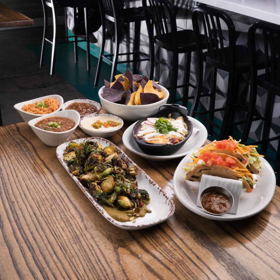 A table full of food including tacos, chips, dips and brussel sprouts