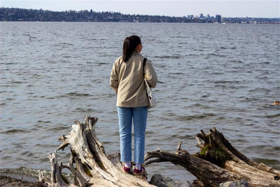 A person wearing jeans and a ponytail stands on a lakeshore facing the water