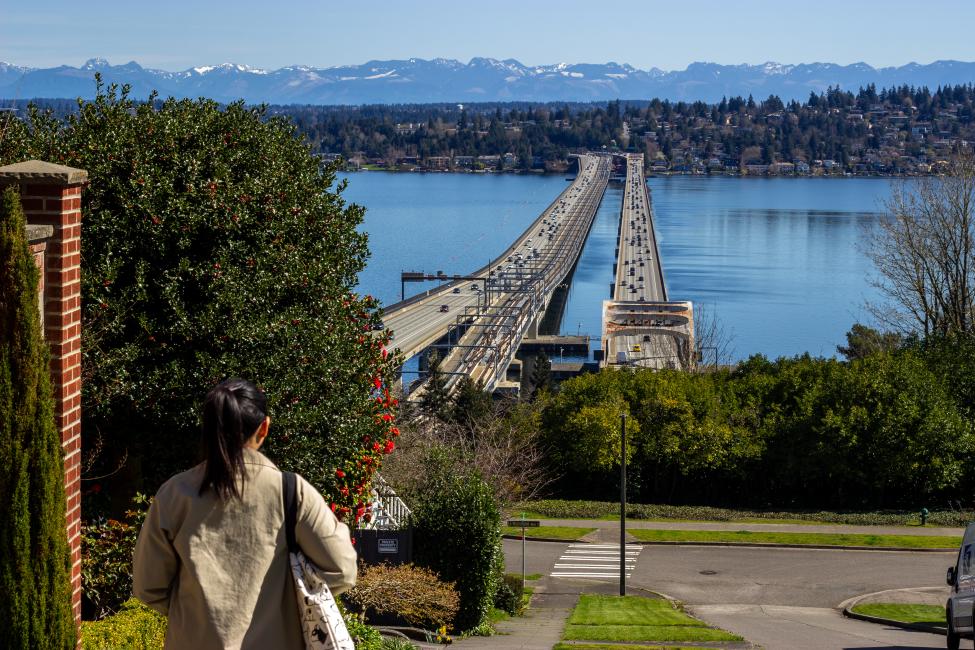 A woman with a ponytail walks on a sidewalk with a view of Lake Washington and the I-90 floating bridges