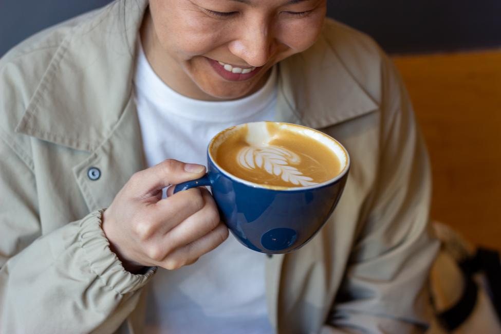 A person wearing a white T-shirt and tan jacket sips coffee from a blue mug