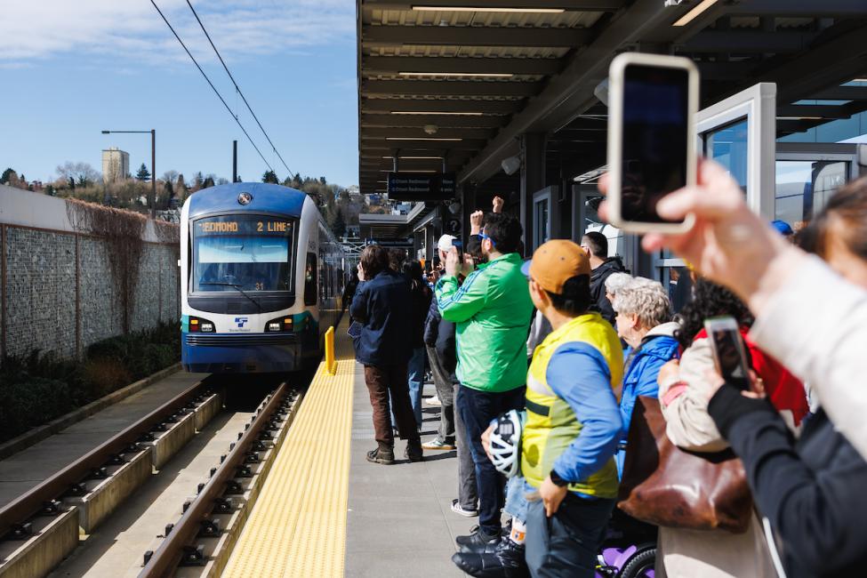 People stand on a platform as a Link train approaches at Judkins Park Station. Many have their phones out to take photos.