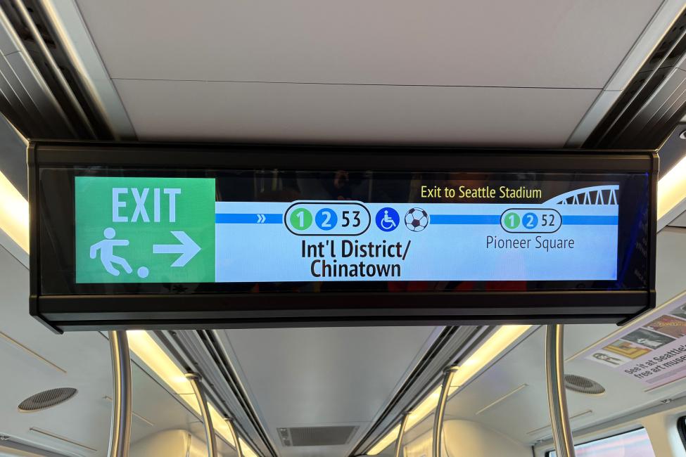 A digital sign onboard a Link train shows the exit for International District Chinatown Station, with icons for the 1 Line, 2 Line and Seattle Stadium above the station name,