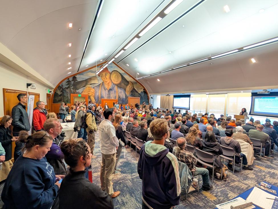 People sit and stand in a large room as a person presents slides about Sound Transit's System Plan