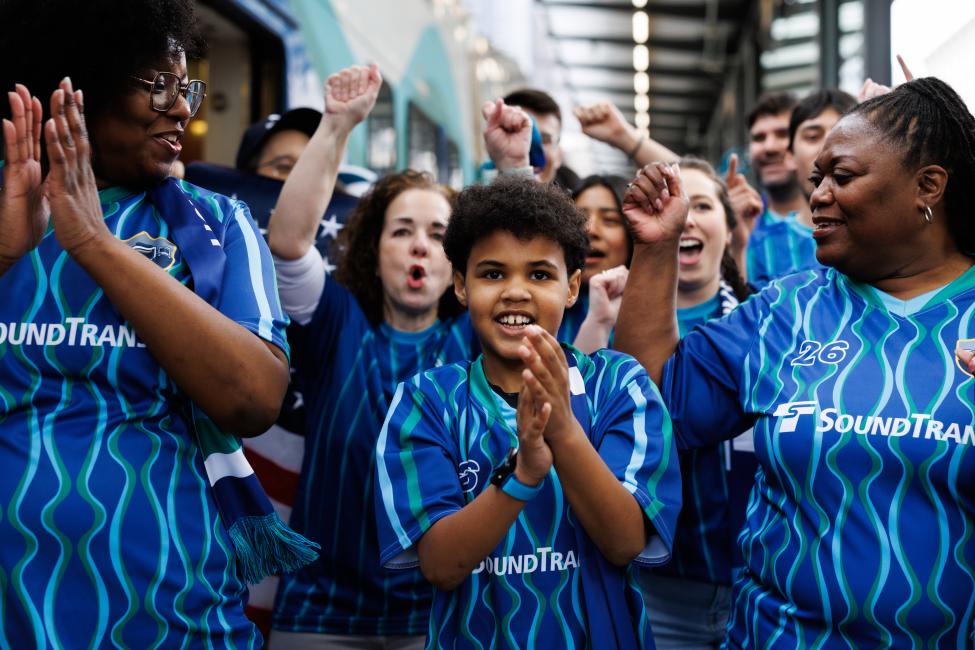A younger person claps his hands together while walking on a train platform among a group of soccer fans. All are wearing blue jerseys.