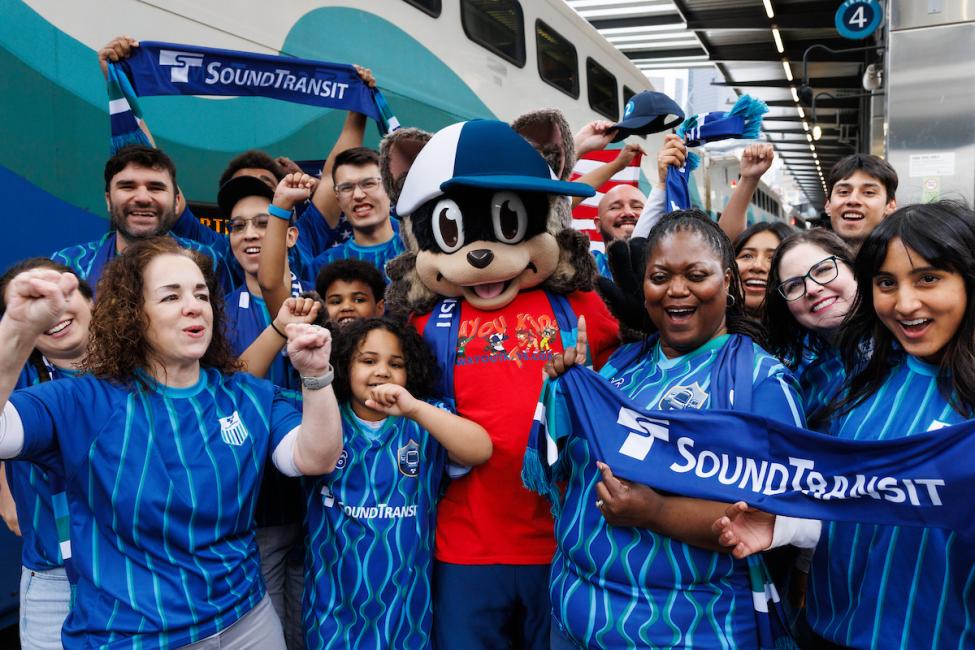 A group of about a dozen soccer fans in blue jerseys on a Sounder platform. Rodney Raccoon, the mascot for WayOut Kids, is in the middle of the group.