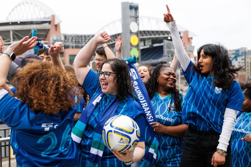 A group of 8-10 fans in blue jerseys cheer and hold a soccer ball in front of Lumen Field