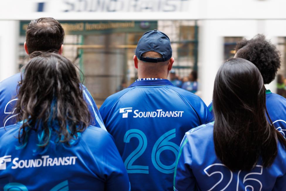 A photo from behind of a group of people wearing Sound Transit merchandise as a train pulls in front of them