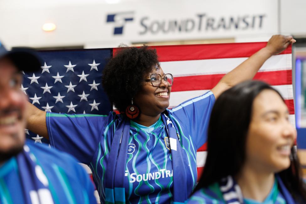 A soccer fan wearing a blue jersey holds a U.S. flag up behind her head. Two other fans are in the foreground of the photo