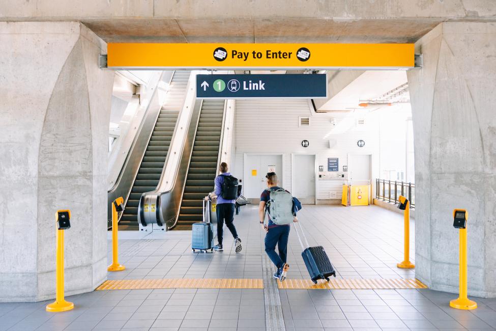 Two people with suitcases walk into the fare paid zone at Northgate Station, delineated by yellow signage. 