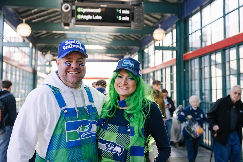 Two people in Seahawks hats and overalls smile by the entrance to International District Chinatown Station