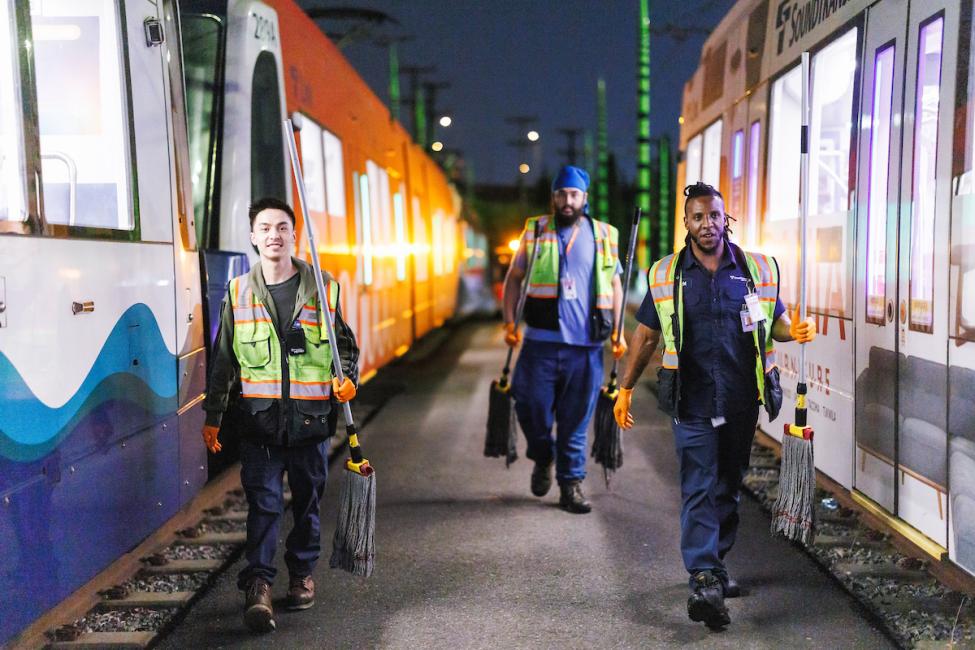 Three workers walk between Link light rail vehicles in a train yard at night