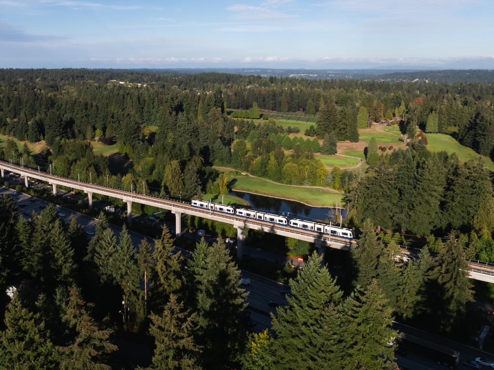 An aerial view of a Link train heading towards Pinehurst Station. There are many trees surrounding the elevated guideway