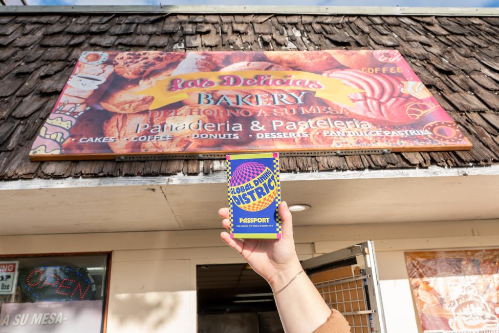 A hand holds up a Global Dining District passport in front of the sign for Las Delicias Bakery