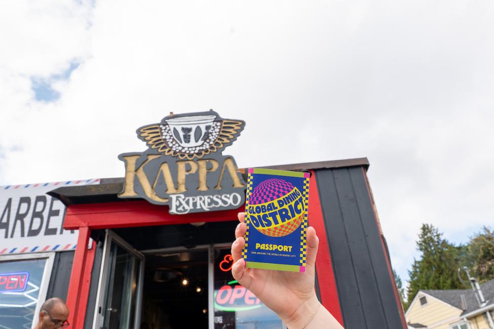 A hand holds up a colorful Global Dining District passport in front of the sign for Kappa Espresso