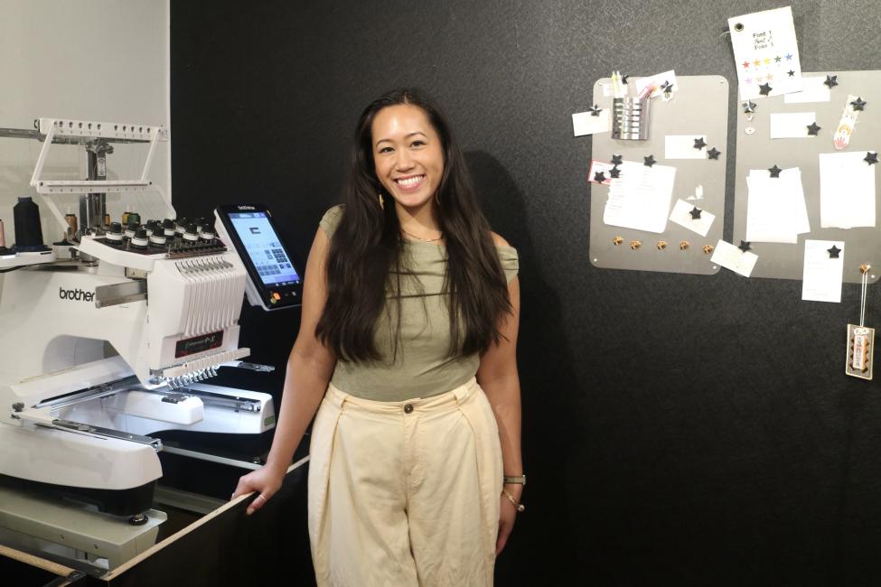 A person with long hair stands in an office with embroidery equipment