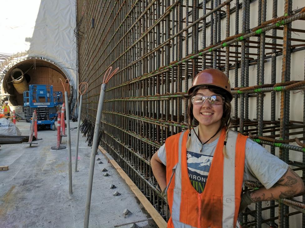 A young construction worker smiles in front of the steel rebar wall she will spend her day connecting with wire. 