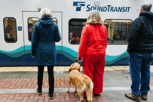 A woman with a cane and a woman with a seeing eye dog wait for the train.