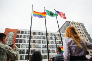 People look up at three flagpoles with the Pride flag, the state of Washington flag and the U.S. flag on display.