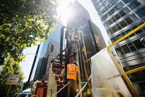 A man in an orange vest stands in front of a drilling rig, with buildings in downtown Seattle in the background.