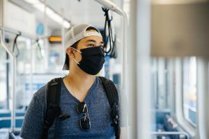 A man wearing a white backwards baseball hat and black face mask looks out the window of a Link train.