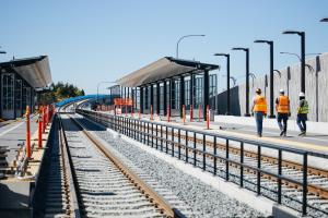 On a sunny day, three construction workers in yellow vests and hard hats walk along the platform at Overlake Village Station, which is still under construction