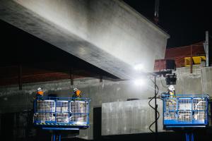 Three construction workers in lifts shine lights on the underside of a large concrete girder. Photo was taken at night.