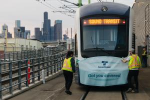 Two construction workers in yellow vests place a sticker that says "We got you covered" on the front of a Link train. The sticker is shaped like a face mask.