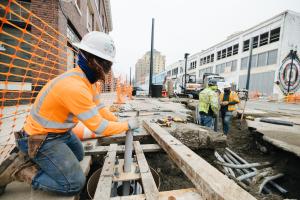 Construction workers wear orange shirts, hard hats and face coverings as they install light rail track in downtown Tacoma.