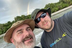 Two men take a selfie on a boat. Both are wearing hats and one is wearing sunglasses.