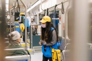 A fare ambassador wearing a blue vest and yellow cap talks to a passenger seated on a light rail train.