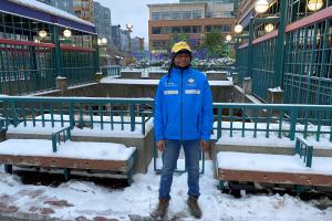 A fare ambassador wearing a blue vest and yellow cap stands outside a light rail station in the snow.