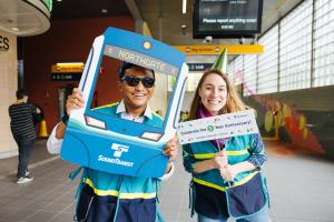 Two Sound Transit employees smile while holding up signs celebrating the one year anniversary of Roosevelt Station.