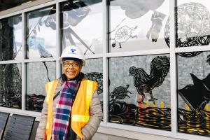 Barbara Earl Thomas wears an orange vest and hard hat while smiling in front of her art at Judkins Park Station.