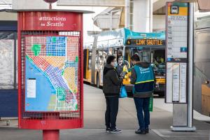 A Sound Transit ambassador in a teal vest talks to a passenger, with a Link shuttle bus in the background.