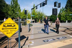 Two people cross the street over light rail tracks in the Rainier Valley, with new safety signage on the pavement reading 'Look'