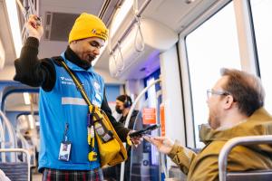A fare ambassador in a blue vest and yellow hat uses a handheld device to perform a fare check with a passenger on a Link train.