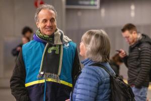 A Sound Transit staff ambassador wears his teal vest and a Sounders scarf while talking to a passenger