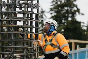 A construction worker in an orange jacket and white hard hat works with rebar