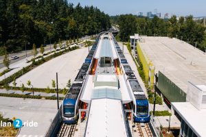 Two trains at South Bellevue Station on a sunny day, with downtown Bellevue in the background. Text reads 'plan 2 ride'