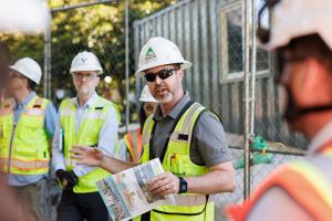 A person wearing a white hard hat and yellow reflective vest talks to a group of people