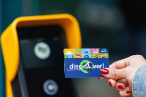 A hand holds an ORCA card up to a yellow tapper. The card says "Discover" and has other designs that commemorate the 1 Line extension to Federal Way