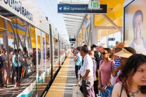 A large group of people wait behind the yellow line on the platform at Lynnwood City Center Station as a Link train pulls up
