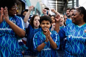 A younger person claps his hands together while walking on a train platform among a group of soccer fans. All are wearing blue jerseys.